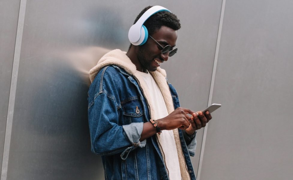 Cool modern smiling african man in wireless headphones listening to music using phone on city street over gray metal wall as background